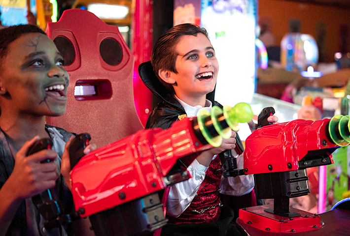 A boy dressed in costume as a zombie plays a shooting game with a boy dressed like a vampire.